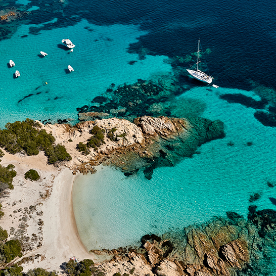 Strand in Sardinien mit Booten im Wasser und einer felsigen Küste im Hintergrund.