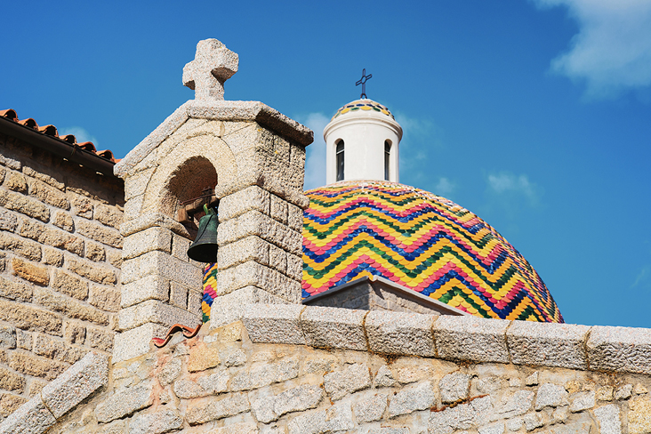 Bunte Kuppel auf einem Gebäude in Sardinien mit einer Glocke oben drauf.