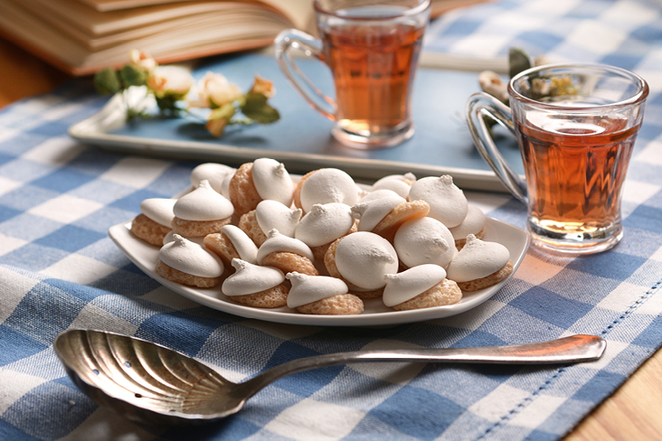 Teller mit weissen Baisers auf einem blau karierten Tischtuch, dazu zwei Tassen Tee und ein geöffnetes Buch – eine Stimmung, die an eine genussvolle Pause in Sizilien erinnert.