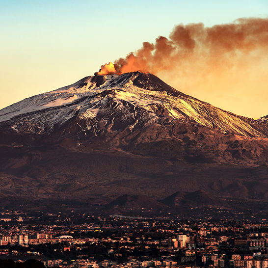 Blick auf den schneebedeckten Etna bei Sonnenuntergang, mit einer Rauchsaeule aus dem Krater und der Stadt am Fuss des Vulkans.