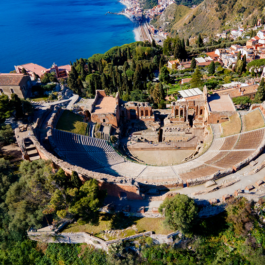 Luftaufnahme des antiken Theaters von Taormina, umgeben von Gruen, mit der Stadt und dem Meer im Hintergrund.