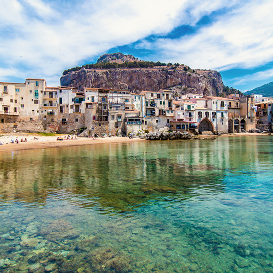 Blick auf Cefalù mit seinen historischen Hausern direkt am Meer, dem belebten Strand und dem markanten Felsen hinter der Stadt unter leicht bewolktem Himmel.