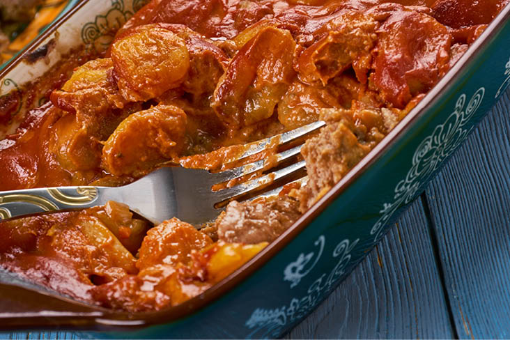 Südafrikanisches Ofengericht mit Kartoffeln, Fleisch und würziger Tomatensauce in einer Keramikform.