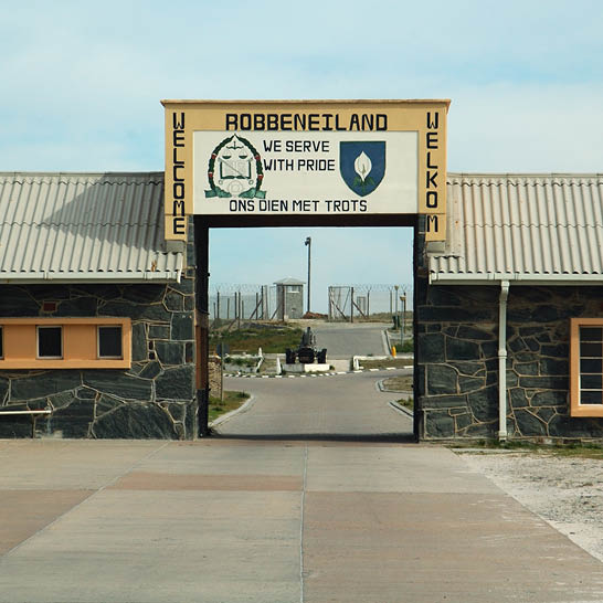 Eingangstor von Robben Island mit Willkommensschild und Blick auf den ehemaligen Gefängnisbereich.