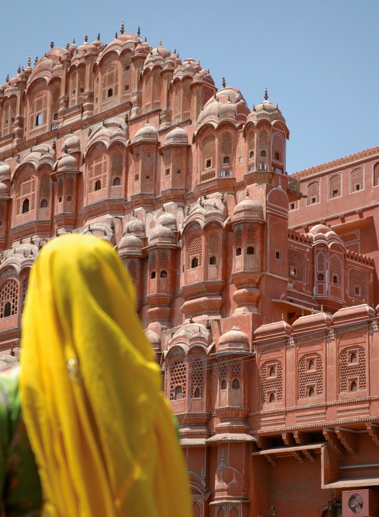 Woman in a yellow Sari and head scarf standing in front of the pink palace walls of Jaipur